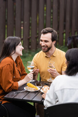 Positive man holding glass of wine near interracial friends and food during picnic