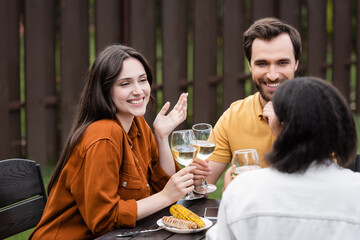 Smiling woman holding wine and talking to multiethnic friends near grilled food and smartphone outdoors