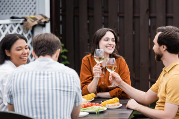 Positive people holding wine while blurred interracial friends talking near tasty food outdoors