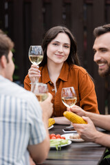 Young woman holding glass of wine and looking at camera near blurred friends during picnic outdoors