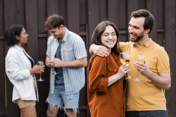cheerful couple holding glasses with wine while hugging near blurred interracial friends on blurred background