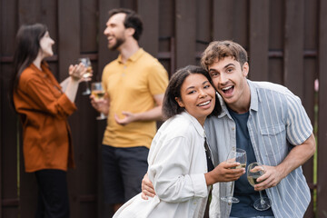 cheerful interracial couple holding glasses with wine near blurred friends on blurred background