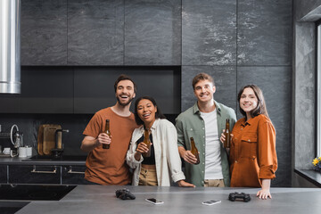 KYIV, UKRAINE - JULY 26, 2022: happy interracial friends holding bottles of beer near joysticks and smartphones on kitchen worktop