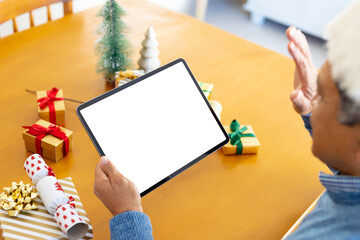 Senior biracial man in santa hat making christmas video call on tablet, waving, copy space on screen