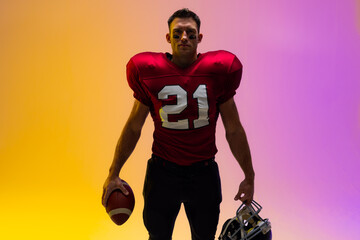 Caucasian male american football player holding helmet and ball with neon yellow and purple lighting