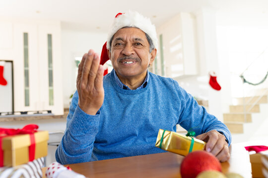Happy senior biracial man in santa hat holding christmas present and gesturing during video call - Powered by Adobe