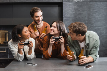 KYIV, UKRAINE - JULY 26, 2022: happy interracial women holding joysticks near male friends with beer in kitchen