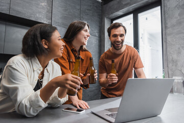 happy multiethnic friends holding bottles of beer near laptop and smartphone with blank screen on kitchen worktop