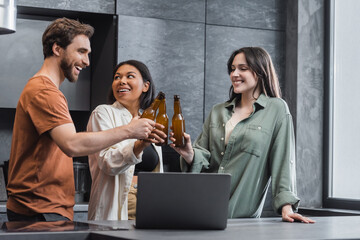 cheerful multiethnic friends holding bottles of beer and clinking near laptop in kitchen