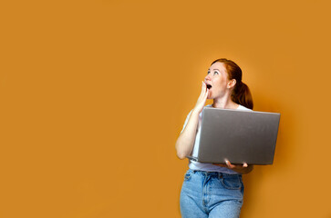 Surprised young red-haired girl in a white empty blank t-shirt posing on a yellow background studio portrait. People lifestyle concept. Copy space for copy. Working on a laptop