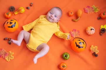 Obraz premium Little child in yellow overall smiles. Newborn baby lies on orange background with decoration pumpkins, autumn leaves and acorns. Top view. Holiday of halloween.