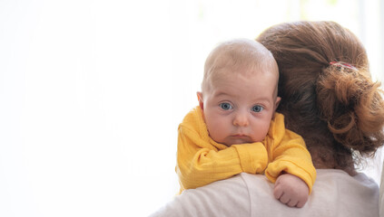 Fototapeta premium Newborn with blue eyes and in yellow t-shirt lies on shoulder by grandma. New worries in family and lifestyle. Concept of child care, feeling safe, parent love.