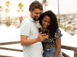 Diverse couple of happy heterosexual lovers looking at smartphone in a city on vacation