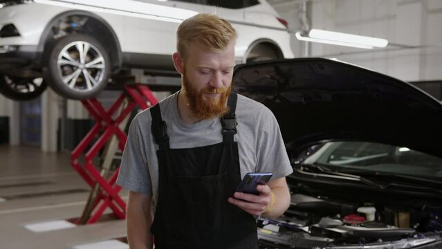 An auto mechanic in a clean uniform with a mobile phone stands against the background of a modern service station - Powered by Adobe