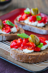 Open bruschetta sandwich with curd cheese, marinated roasted peppers and grilled olives on a red ceramic plate on a wooden background. Sandwich recipes. Antipasti.