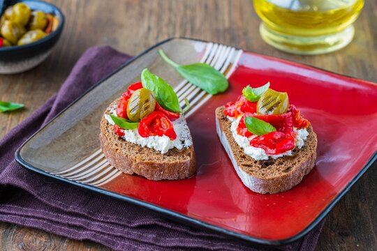 Open bruschetta sandwich with curd cheese, marinated roasted peppers and grilled olives on a red ceramic plate on a wooden background. Sandwich recipes. Antipasti.
