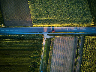 Aerial drone view looking down on a rural road with a car driving along it. On a bright sunny day, farmland and crops can be seen either side of the road. Sunset with long shadows
