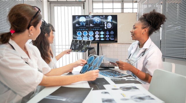 Diverse group female doctors meeting looking at medical x-ray then diagnosis and discussing before surgery at hospital. Surgeon and radiologist teamwork talking, analysis x-ray film before treatment. - Powered by Adobe