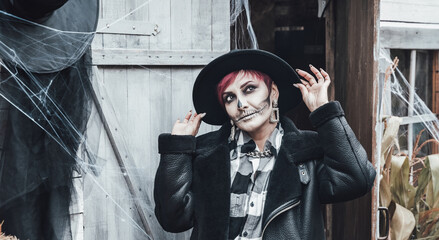 Scary woman, girl celebrating halloween.Terrifying black skull half-face skeleton makeup and witch costume in stylish image. Jacket, hat.Horror,fun at photoshoot, holiday party near barn on street