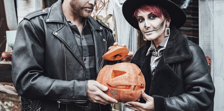 Scary love family couple man,woman celebrating halloween with pumpkin jack-o-lantern.Terrifying black skull half-face makeup,witch costumes,stylish images,jacket,hat. Fun,holiday party at street barn