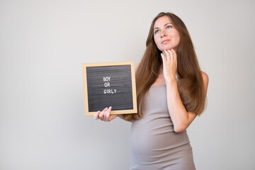 Attractive caucasian pregnant thoughtful dreaming woman holding board with boy or girl question on...