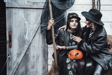 Scary family,mother,daughter celebrating halloween. Street barn. Pumpkin jack-o-lantern.Terrifying black skull half-face makeup and witch costumes, broom,stylish images hat, jackets. Children's party