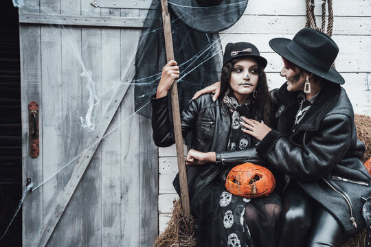 Scary family,mother,daughter celebrating halloween. Street barn. Pumpkin jack-o-lantern.Terrifying black skull half-face makeup and witch costumes, broom,stylish images hat, jackets. Children's party