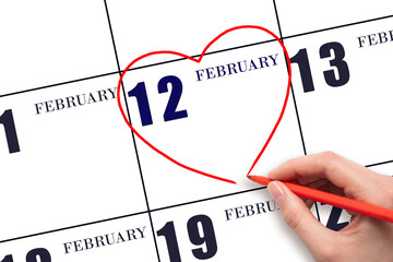 A woman's hand drawing a red heart shape on the calendar date of 12 February. Heart as a symbol of love.