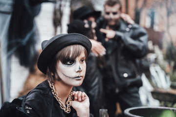 Beautiful scary little girl celebrating halloween. Terrifying black, white half-face makeup and witch costume, stylish image. Horror, fun at children's party in barn on street. Hat, fur coat, chain