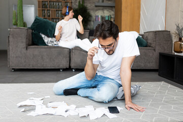 Young husband sitting on the floor with many bills and checks and calculating expenses of his spender wife who sitting on the couch and relaxing