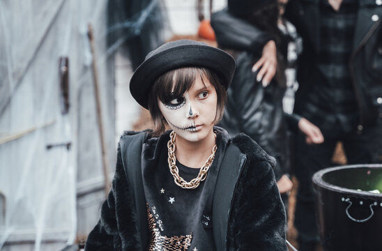 Beautiful scary little girl celebrating halloween. Terrifying black, white half-face makeup and witch costume, stylish image. Horror, fun at children's party in barn on street. Hat, fur coat, chain