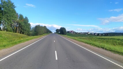An asphalt road with markings and sandy shoulders passes among grassy meadows. The road is flanked by trees. In the distance is a village. Sunny weather and blue skies with clouds