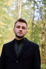 Handsome guy in a black coat in the forest. Cute guy with stubble and a model hairstyle is posing in the forest while standing in a coat.