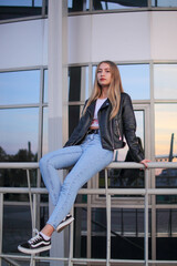 a young beautiful blonde girl is sitting on the railing near a new modern building in a leather jacket and jeans in autumn