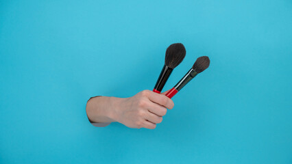 A woman is holding two large brushes for cosmetics. Woman's hand sticking out of blue paper background. 