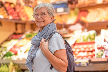 Attractive senior woman with backpack and scarf walking for shopping at the farmer market looking for fresh fruit and vegetables