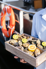 A variety of fresh oysters with lime and lemon in wooden box. Fresh seafood. Outdoor cafe terrace. Blurred background with a view of the yacht club.