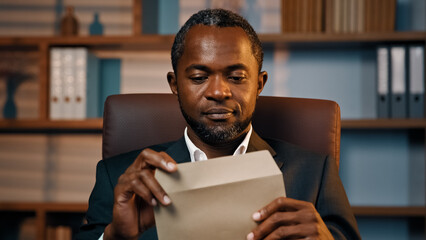 Successful businessman sits in office opening paper envelope reading letter with good news received...