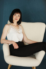 attractive business woman sits in comfy white chair. Smiling brunette lady with short hair wears black office trousers and white blouse, high-heels isolated on blue green background. copy space.