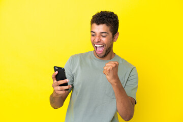 Young handsome Brazilian man isolated on yellow background with phone in victory position