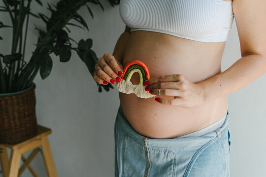 A pregnant girl in a denim jumpsuit holds a rainbow in her hands for the decor of a children's room. Great time to have a baby. Future mom. Rainbow macrame.