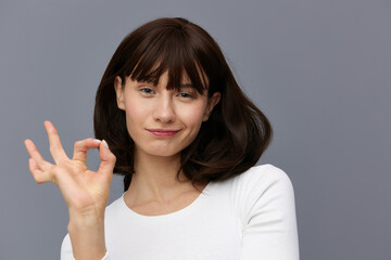  beautiful, happy woman smiles pleasantly looking at the camera standing on a gray background in a white T-shirt and shows the ok sign near her face. Horizontal studio photography with empty space
