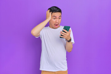 Shocked young Asian man in white t-shirt looking at smartphone with wow expression isolated on purple background