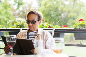 Senior woman wearing sunglasses reading a menu while sitting on the terrace of the restaurant.