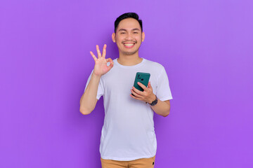Smiling young Asian man in white t-shirt using smartphone and showing ok gesture isolated on purple background