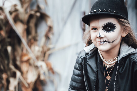 Beautiful scary little girl celebrating halloween. Terrifying black, white half-face makeup,witch costume, stylish image. Horror, fun at children's party in barn on street. Hat, jacket