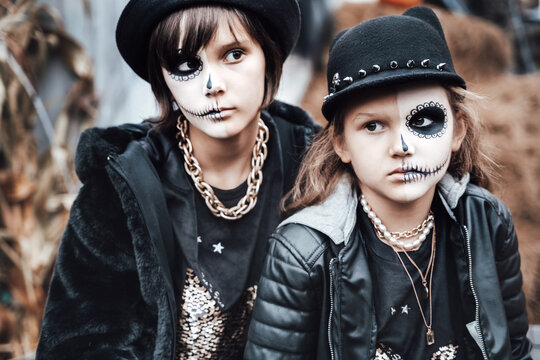 Beautiful scary little girls celebrating halloween. Terrifying black, white half-face makeup,witch costume, stylish image. Horror, fun at children's party in barn on street. Hat, jacket