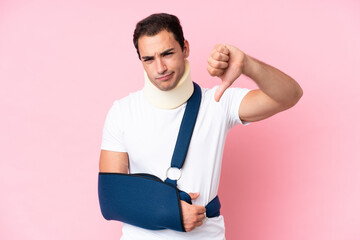 Young caucasian man with broken arm and wearing a sling isolated on pink background showing thumb...