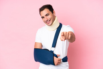 Young caucasian man with broken arm and wearing a sling isolated on pink background pointing front...
