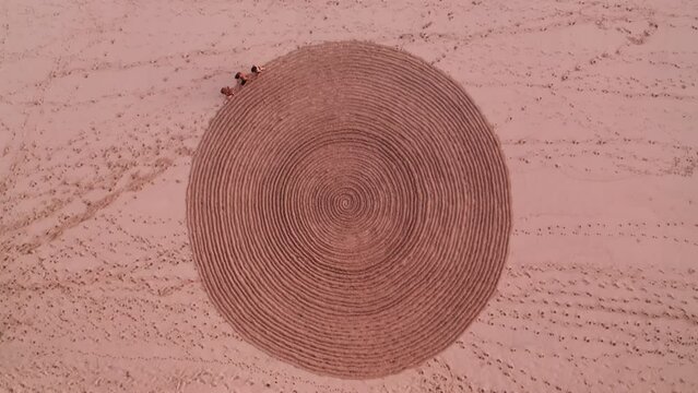 Aerial top view of people trampling down mysterious spiral circus on sand. Fake mystery and sports exercise for tourists on seaside. Rite with ancient symbol. Enigmatic sign on sandy beach in summer.
